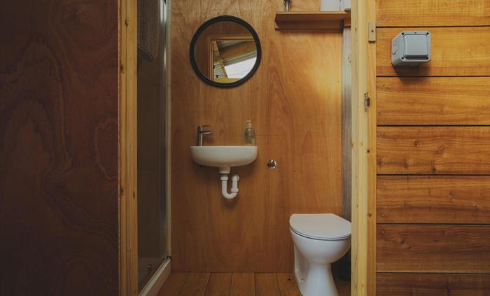 Looking in to a bathroom made of plywood.  There is a warm glow to the image. A shower is seen to the right and in the centre is a basin with mirror a